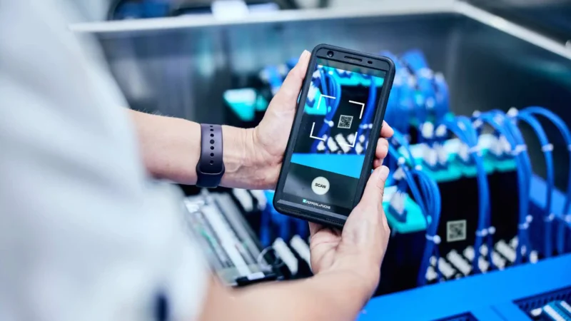Headerbild zeigt ein Mobil Gerät auf dem ein QR Code gescannt wird. Im Hintergrund eine undeutlich gemachte blaue Maschine auf dem der QR Code angebracht ist.