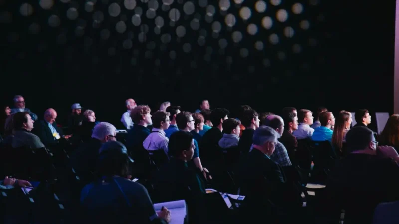 Schmuckbild: Konferenz: Menschen sitzen auf Stühlen in einem abgedunkeltem Raum und schauen nach vorne auf eine Tribüne. Einzelne Personen schreiben sich Notizen auf .
